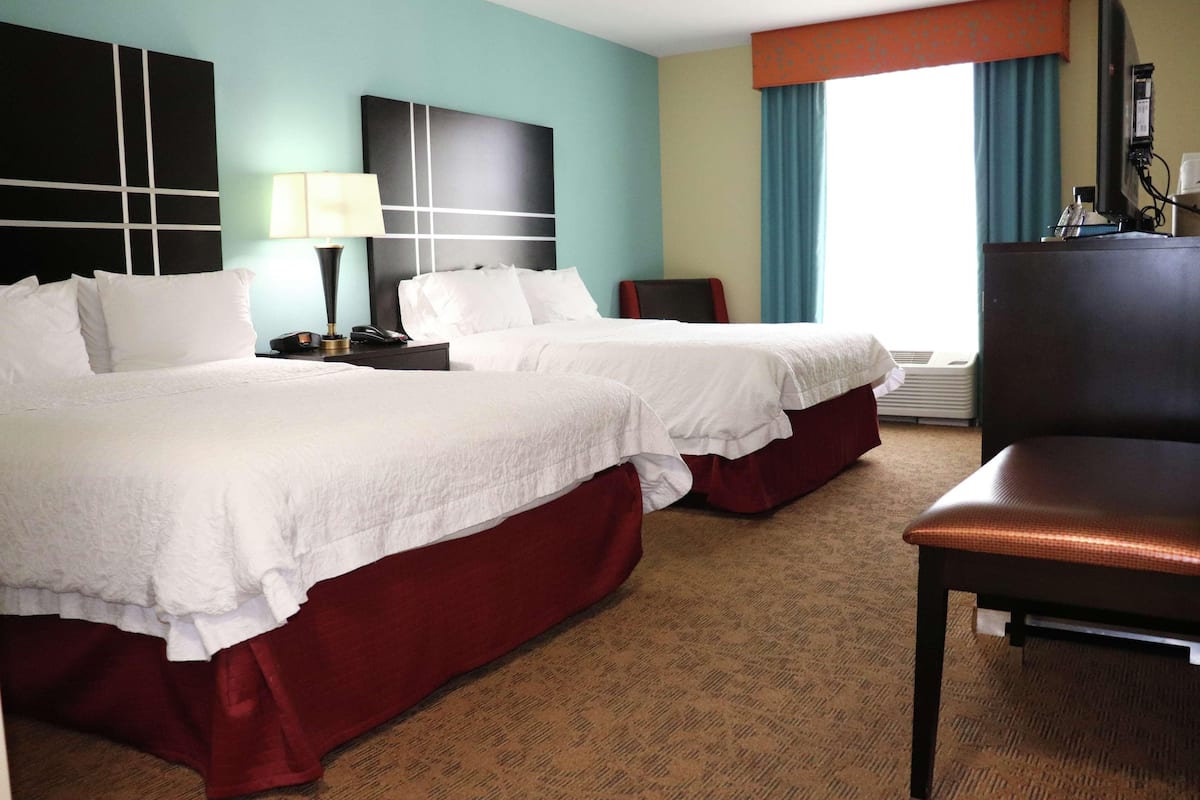 A hotel room with two beds, a desk, a chair, and a large window with curtains.