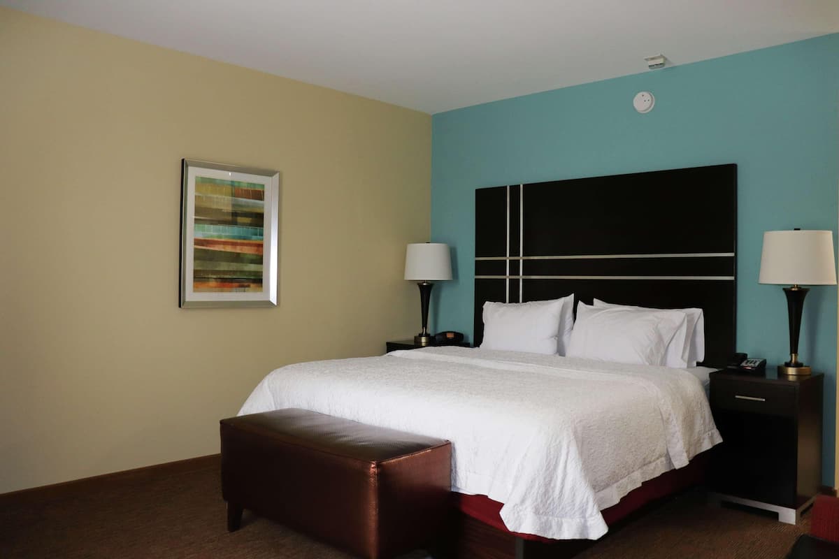 A hotel room with a bed, a nightstand, a lamp, a bench, and a wall art piece.