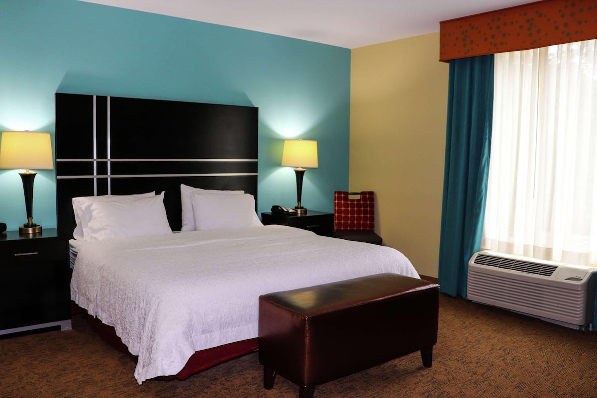 A hotel room with a large bed, two bedside lamps, a bench, an air conditioning unit, and a window with curtains.