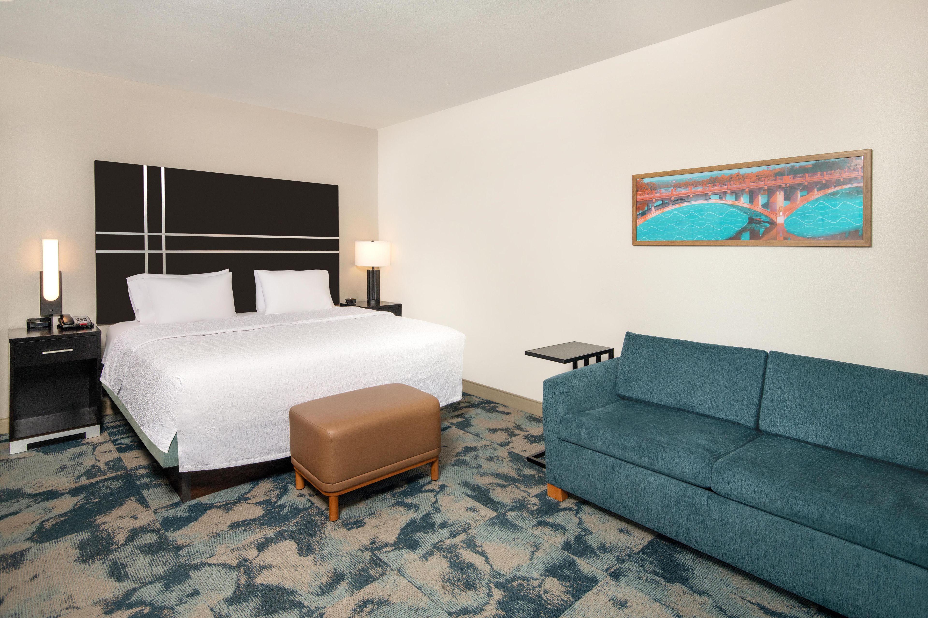 A hotel room with a bed, bedside tables, a nightstand, a sofa, and a painting on the wall.