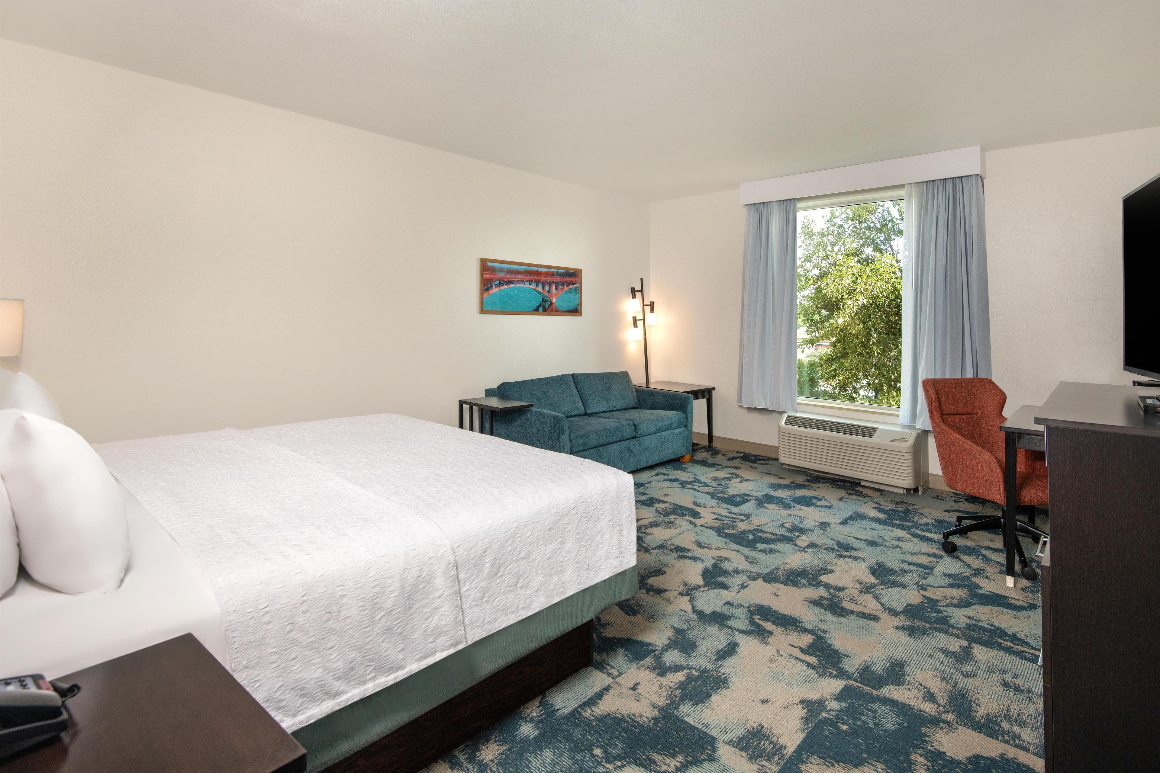 A hotel room with a bed, a desk, a chair, a lamp, and a painting on the wall.