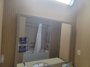 A bathroom with a large mirror, a shower, a towel rack, and a hand sanitizer dispenser. - Hampton Inn Austin/Oak Hill (Austin)