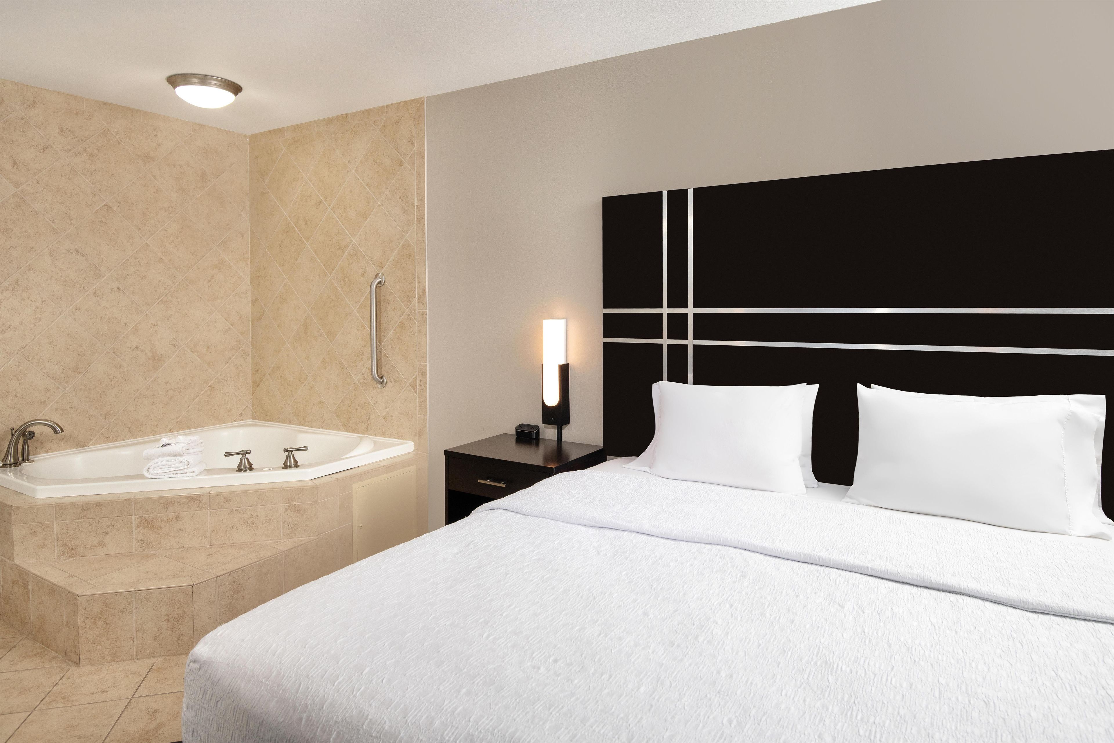 Habitación de hotel con una cama grande, una bañera independiente y una mesita de noche con lámpara.