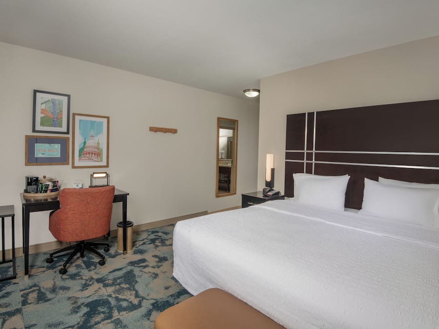 Habitación de hotel con una cama grande, un escritorio con silla, un bote de basura pequeño y cuadros en la pared.