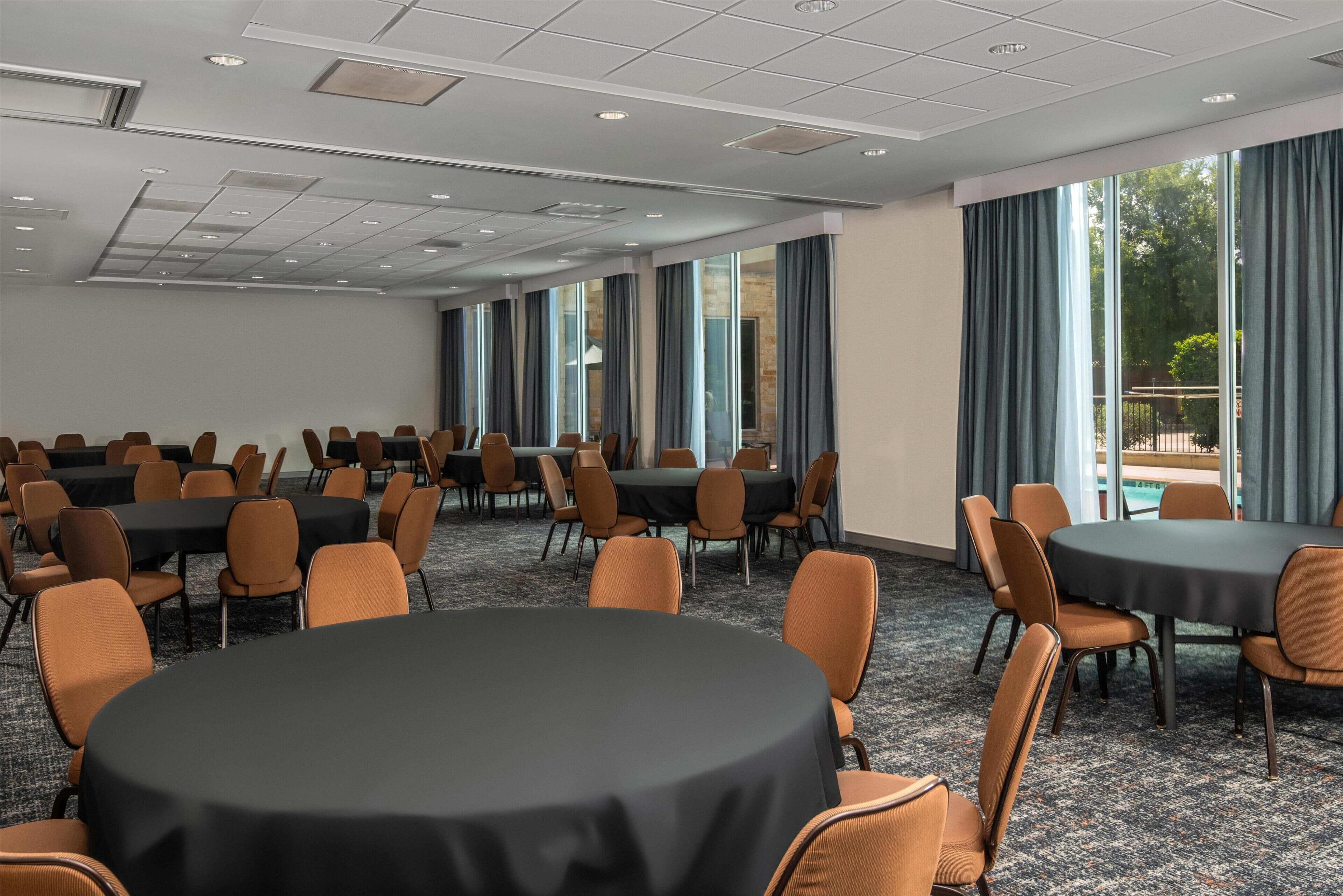 Un salón de conferencias con mesas y sillas redondas, ventanas amplias y vistas al exterior.