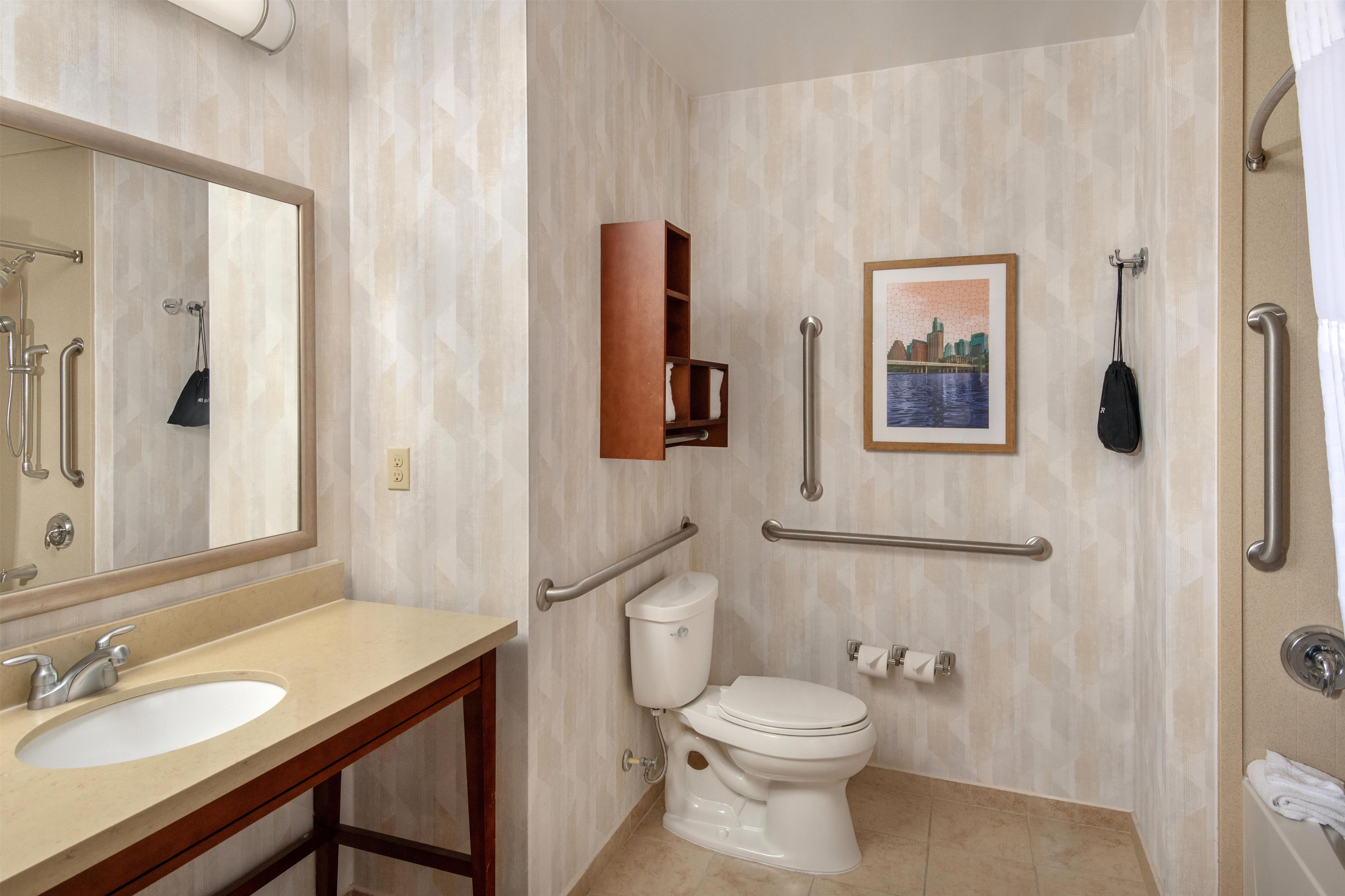 Baño con lavamanos, inodoro y espejo. Se observan barras de apoyo y un perchero para toallas.