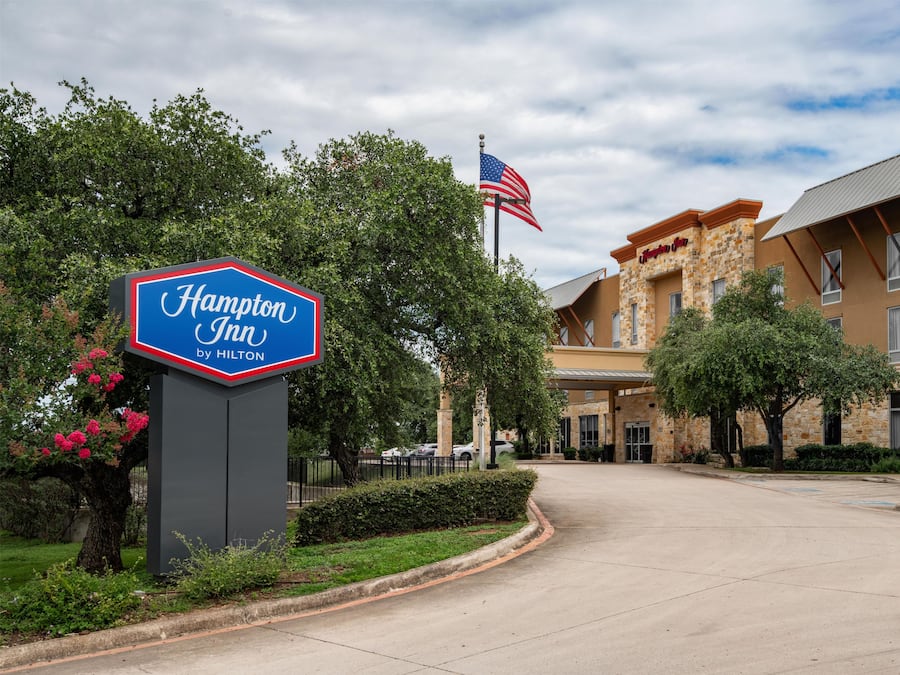 Placa de um Hampton Inn by Hilton com uma bandeira americana, cercada por vegetação e um edifício de fachada de pedra e várias janelas.