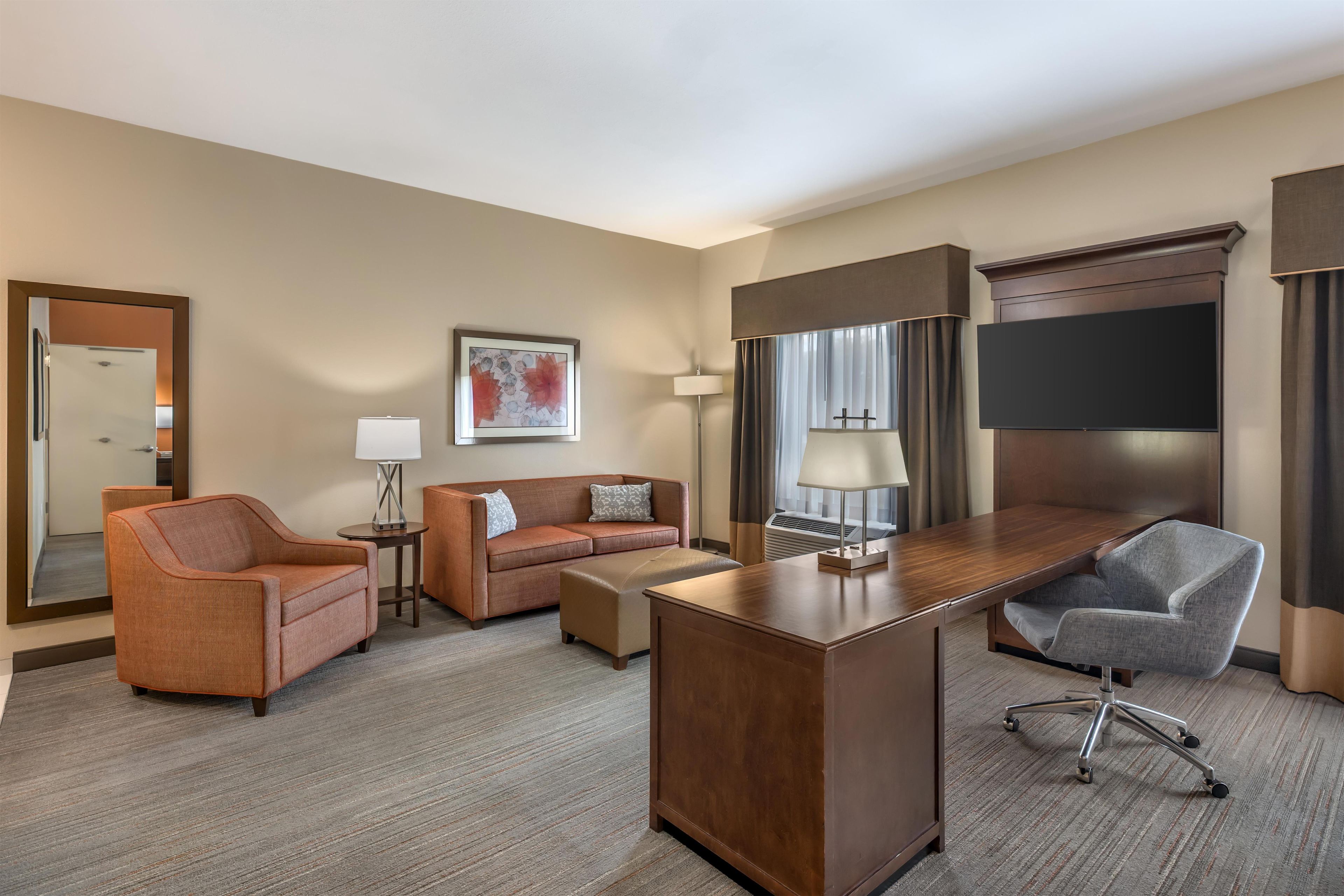 A hotel room with a desk, two armchairs, a sofa, a flat-screen TV, a lamp, and a framed picture on the wall.