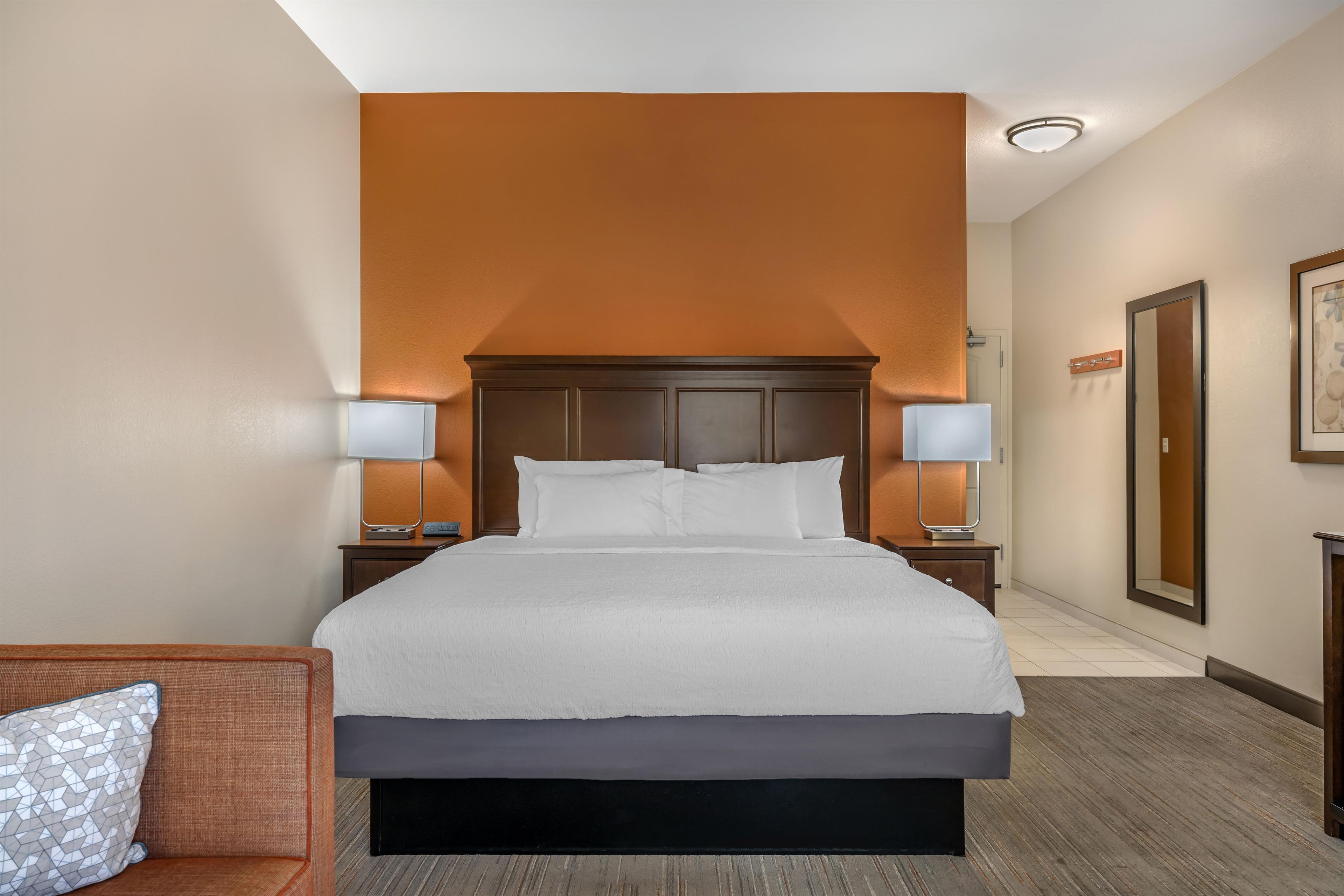 A hotel room with a large bed, two bedside tables with lamps, a nightstand, a wooden headboard, and a framed picture on the wall.