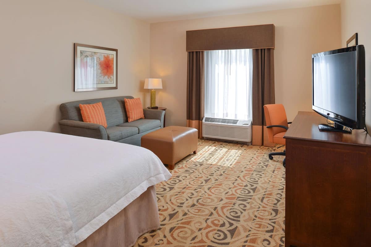 A hotel room with a large bed, a sofa, an ottoman, a desk with a TV, and a window with curtains.