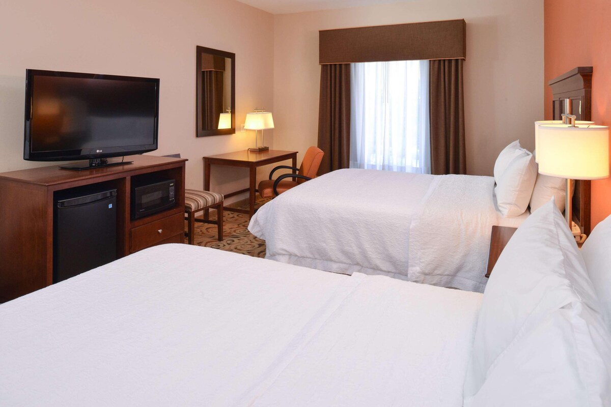 A hotel room with two beds, a television, a desk with a lamp, and a window with curtains.