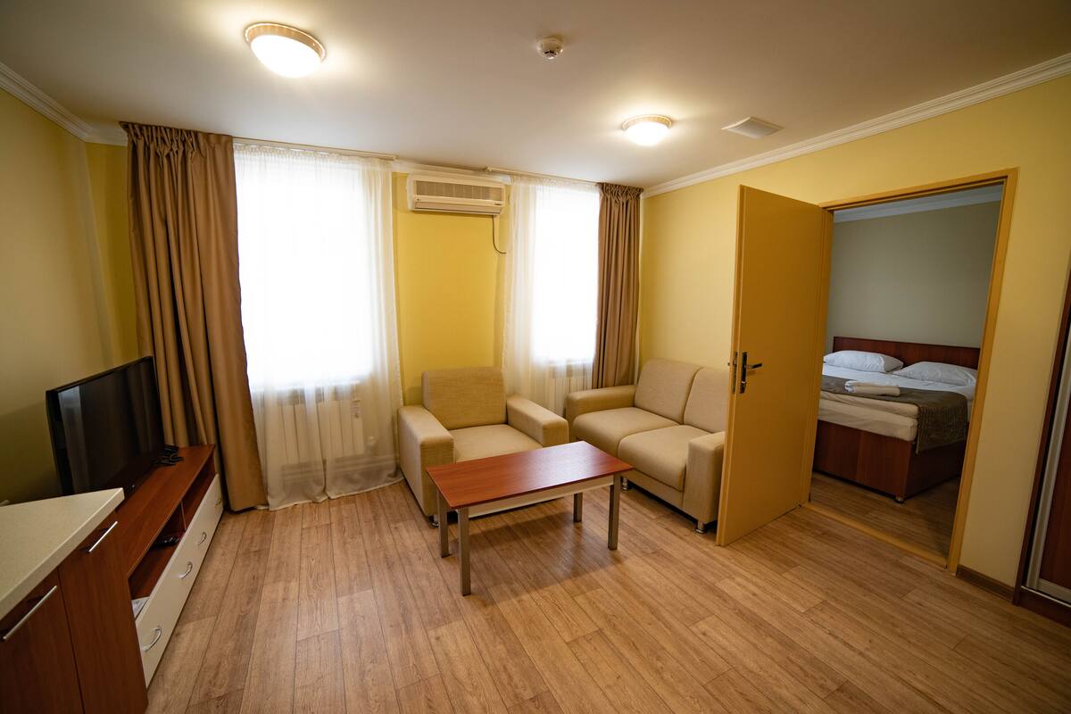 Habitación de hotel con piso de madera, sofás color beige, una mesa de centro de madera, televisor y una cama con cabecera de madera.