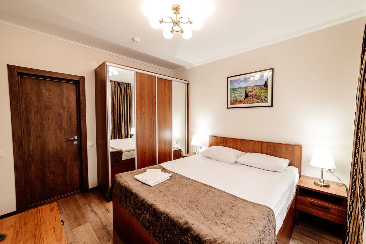Una habitación de hotel con una cama de madera, un armario, un espejo, una mesita de noche con una lámpara y un cuadro en la pared.