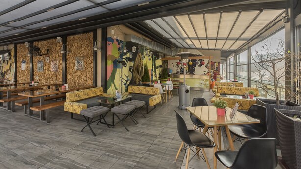 A modern restaurant with a high ceiling, glass walls, and a variety of seating options including tables, benches, and cushioned seating. - Hotel Lirak (Tetovo)