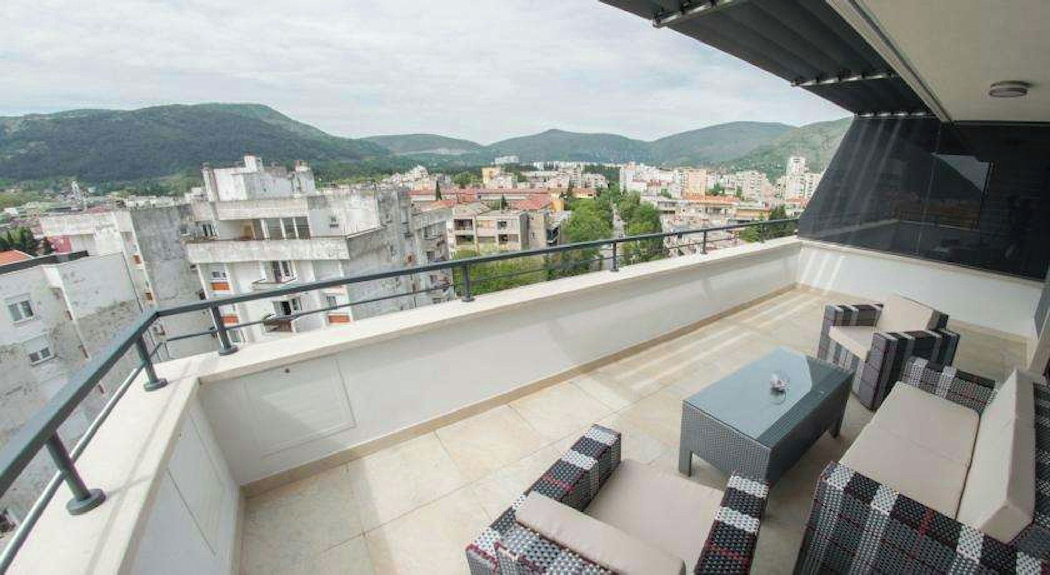 A balcony with outdoor furniture, a view of a cityscape, and mountains in the distance.