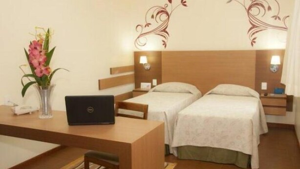 A hotel room with two beds, a desk with a laptop, a vase with flowers, and decorative wall art. - Hotel Premium Pirassununga (Pirassununga)