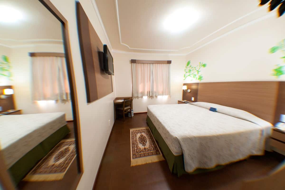 A hotel room with a bed, a desk, a chair, a small table, a nightstand, a mirror, and a window with curtains.