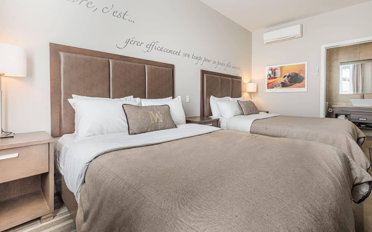A hotel room with two beds, a nightstand, a lamp, a mirror, and a wall with a French phrase.