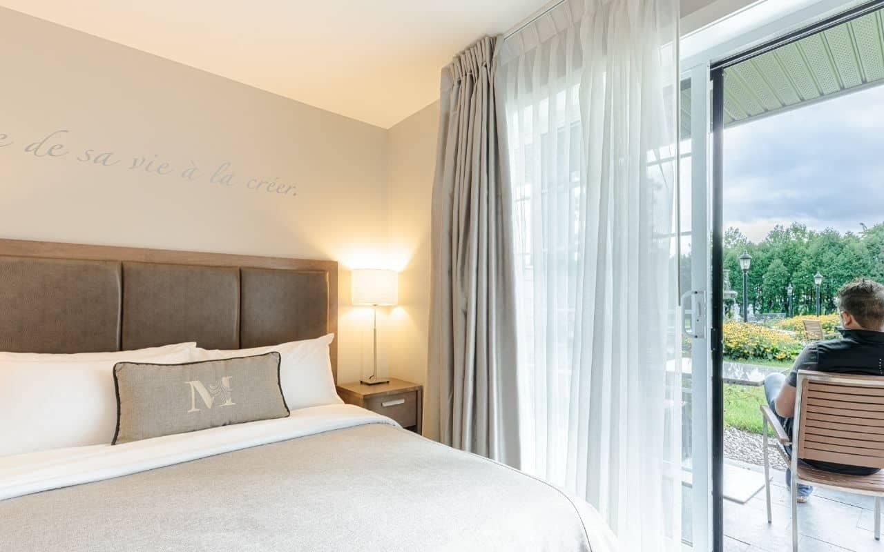 Et hotelværelse med en stor seng, et natbord med en lampe og udsigt til en have gennem en glasdør.