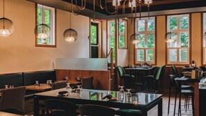 Un restaurante con mesas y sillas de madera, lámparas colgantes y amplios ventanales con vistas a zonas verdes.