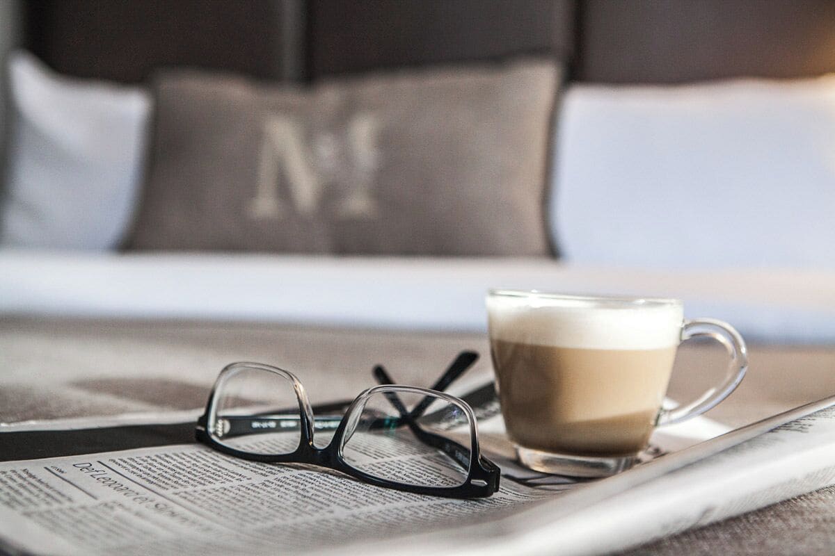 Ein Bett mit einem Kissen mit einem großen 'M', eine Kaffeetasse und eine Brille auf einer Zeitung.