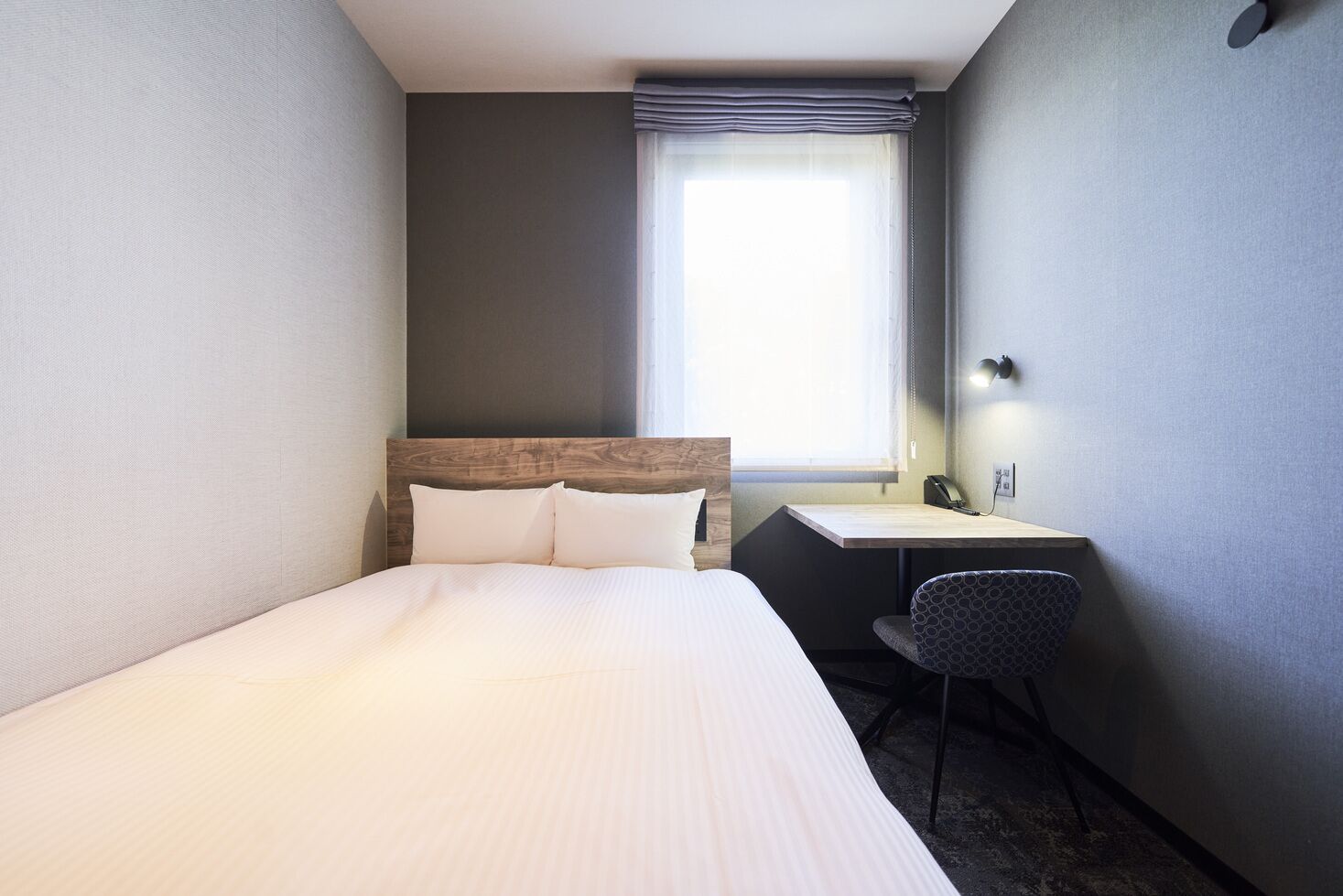 Ein Hotelzimmer mit einem Bett, einem Schreibtisch, einem Stuhl, einem fensterbündigen Fenster mit Vorhängen und einem an der Wand befestigten Telefon.