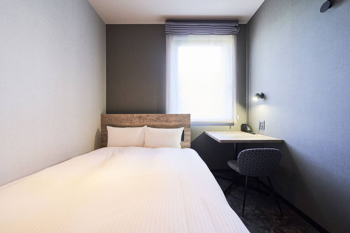 Ein Hotelzimmer mit einem Bett, einem Schreibtisch, einem Stuhl, einem fensterbündigen Fenster mit Vorhängen und einem an der Wand befestigten Telefon.