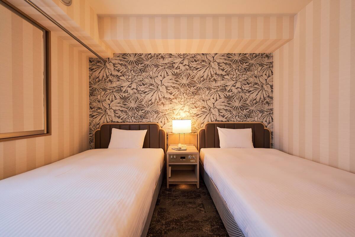 Ein Hotelzimmer mit zwei Einzelbetten, einem Nachttisch mit Lampe und blumengemustertem Wandpapier.