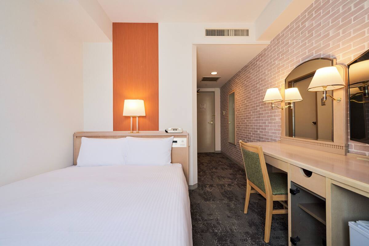 Ein Hotelzimmer mit Bett, Schreibtisch mit Stuhl, Spiegel und Wandlampe.