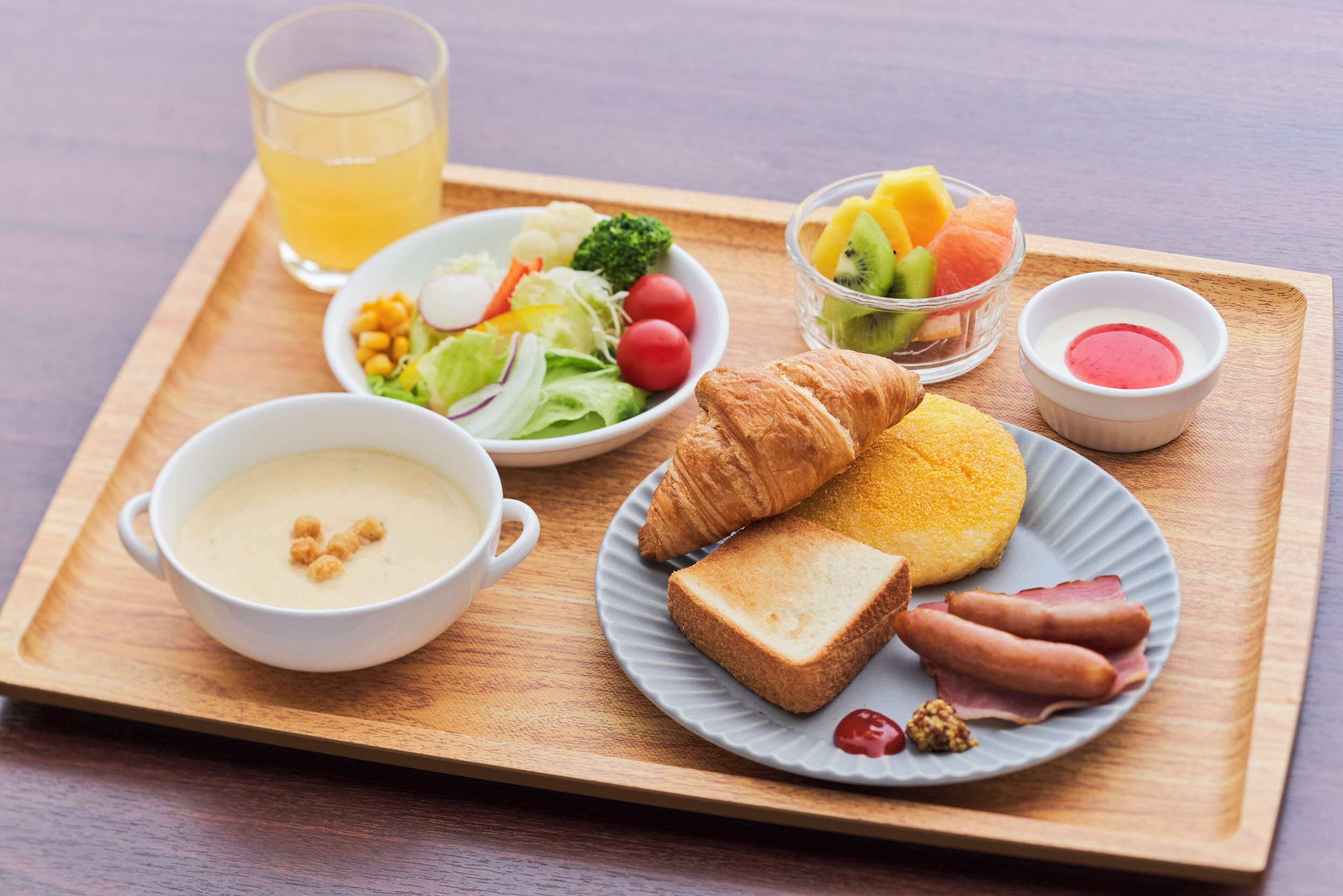 A breakfast tray with croissants, ham, toast, soup, salad, fruit, and a glass of juice.