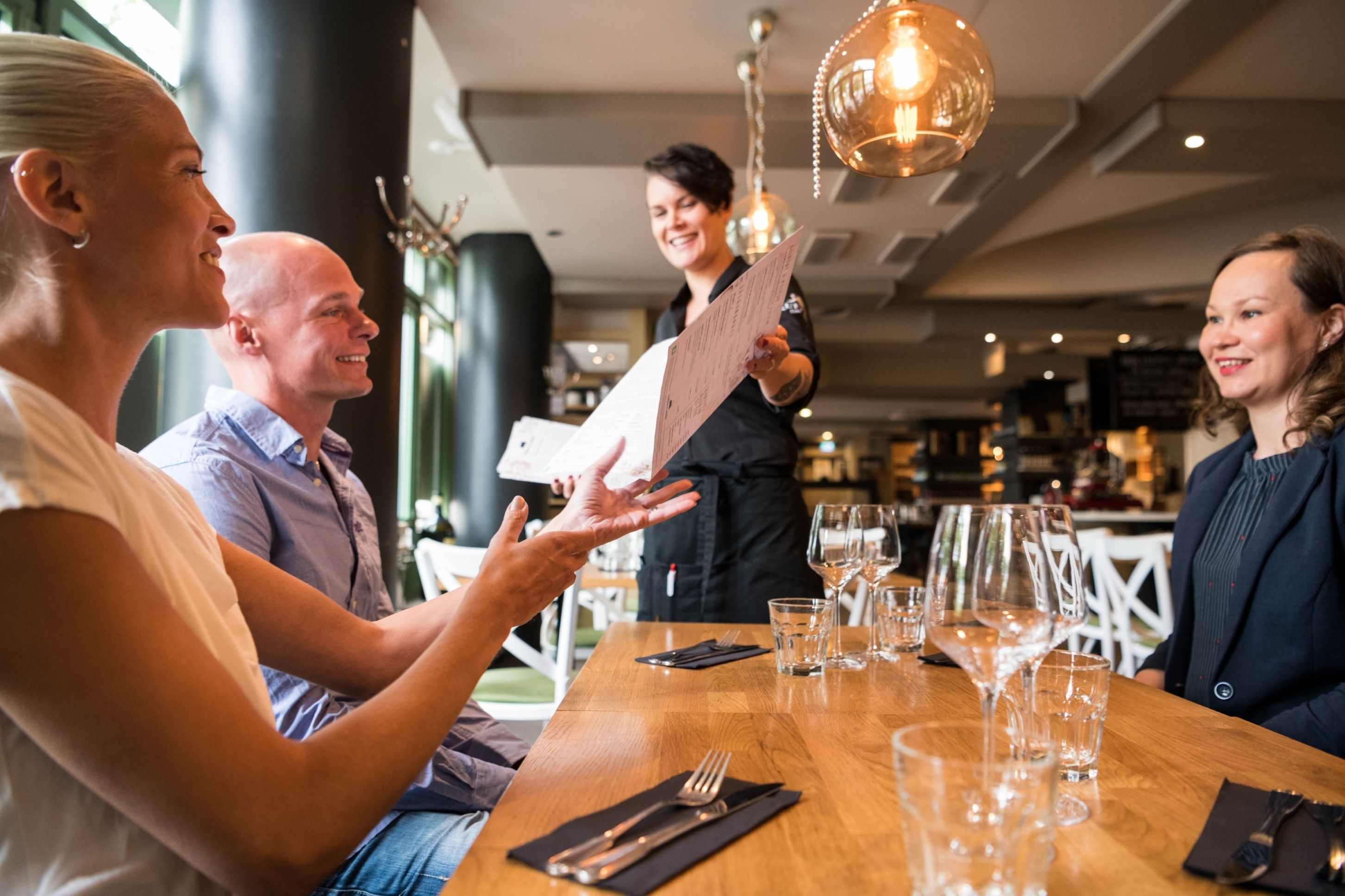A restaurant setting with a wooden table, wine glasses, and a menu being held by a server.