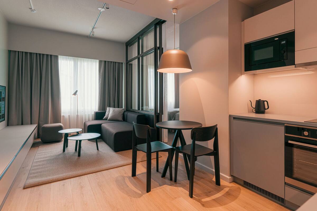 Un espace de vie moderne et compact comprenant un canapé, deux petites tables et des chaises, une kitchenette équipée d’un micro-ondes et d’une bouilloire, ainsi que de grandes fenêtres avec des rideaux.