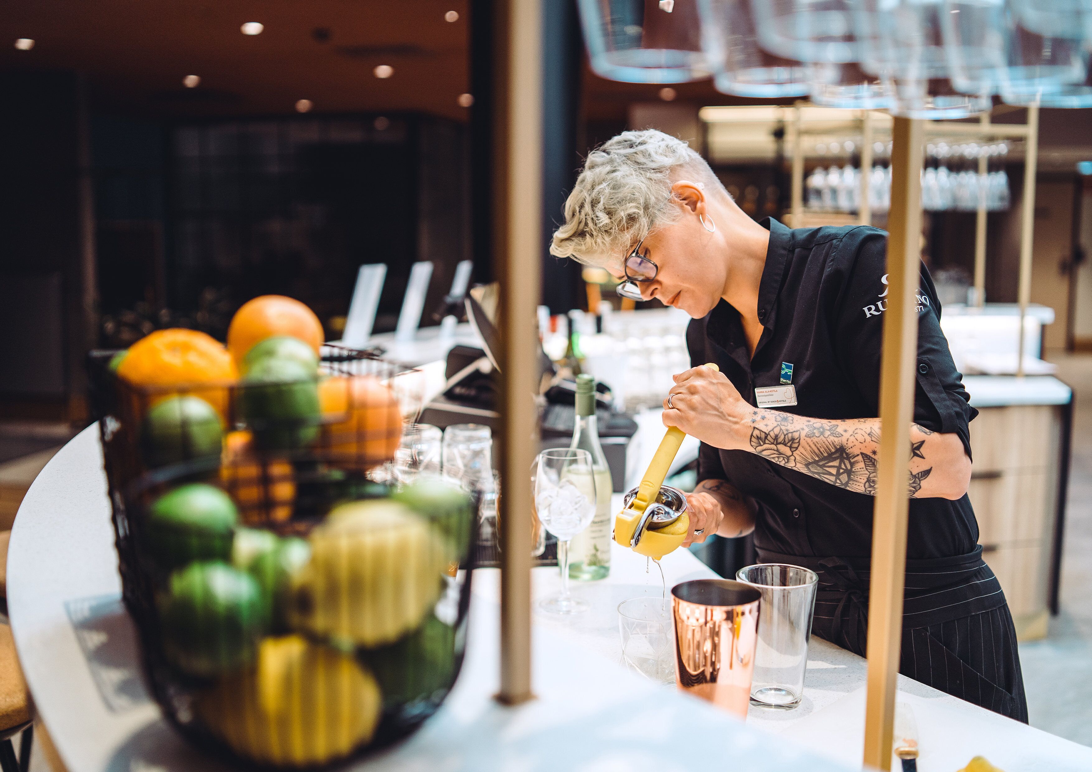 A bartender preparing a drink at a bar with a variety of fruits and bottles in the background.