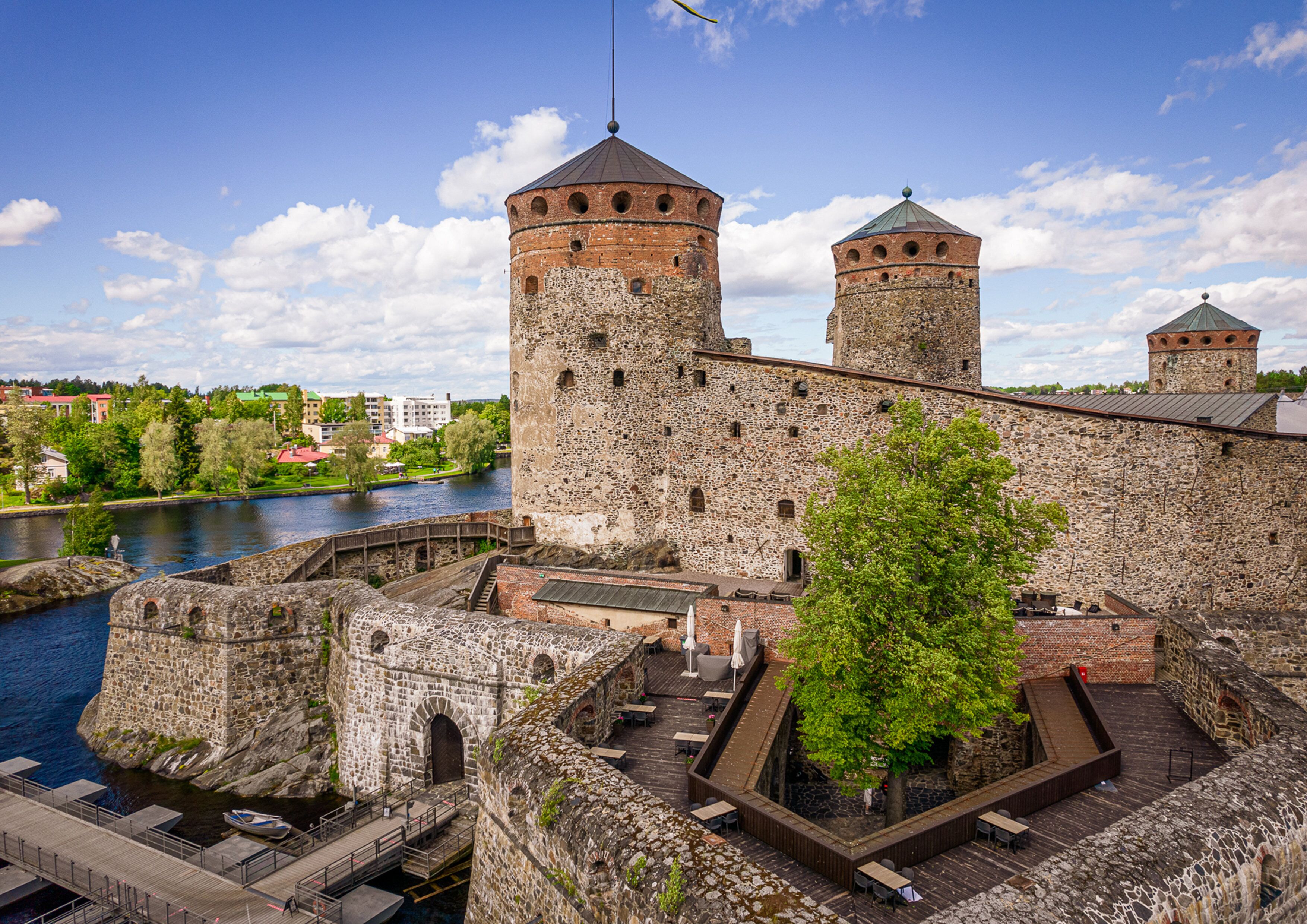 A historic stone castle with two towers, a river in the background, and a wooden deck with outdoor seating.