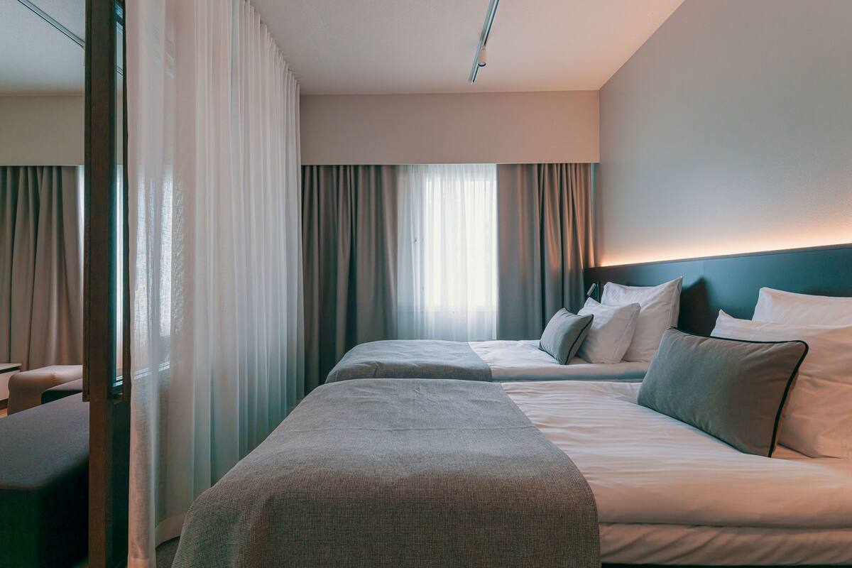 Une chambre d’hôtel avec deux lits, une grande fenêtre avec des rideaux, une tête de lit et une table de chevet.