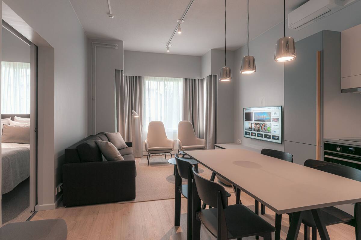 Une chambre d’hôtel moderne équipée d’un canapé, d’une table à manger et de chaises. Un téléviseur est fixé au mur, et un coin cuisine comprend des placards et un four à micro-ondes.