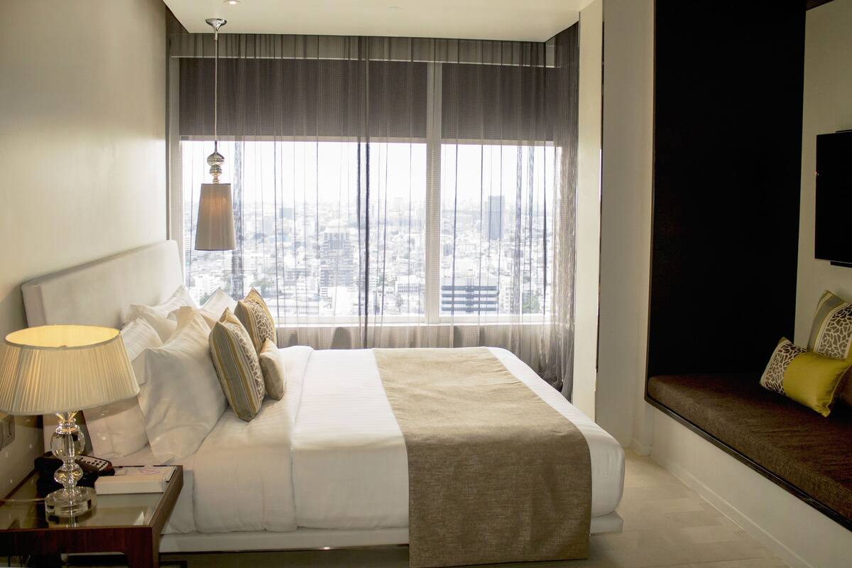 A hotel room with a large bed, a nightstand with a lamp, and a view of the city through large windows.