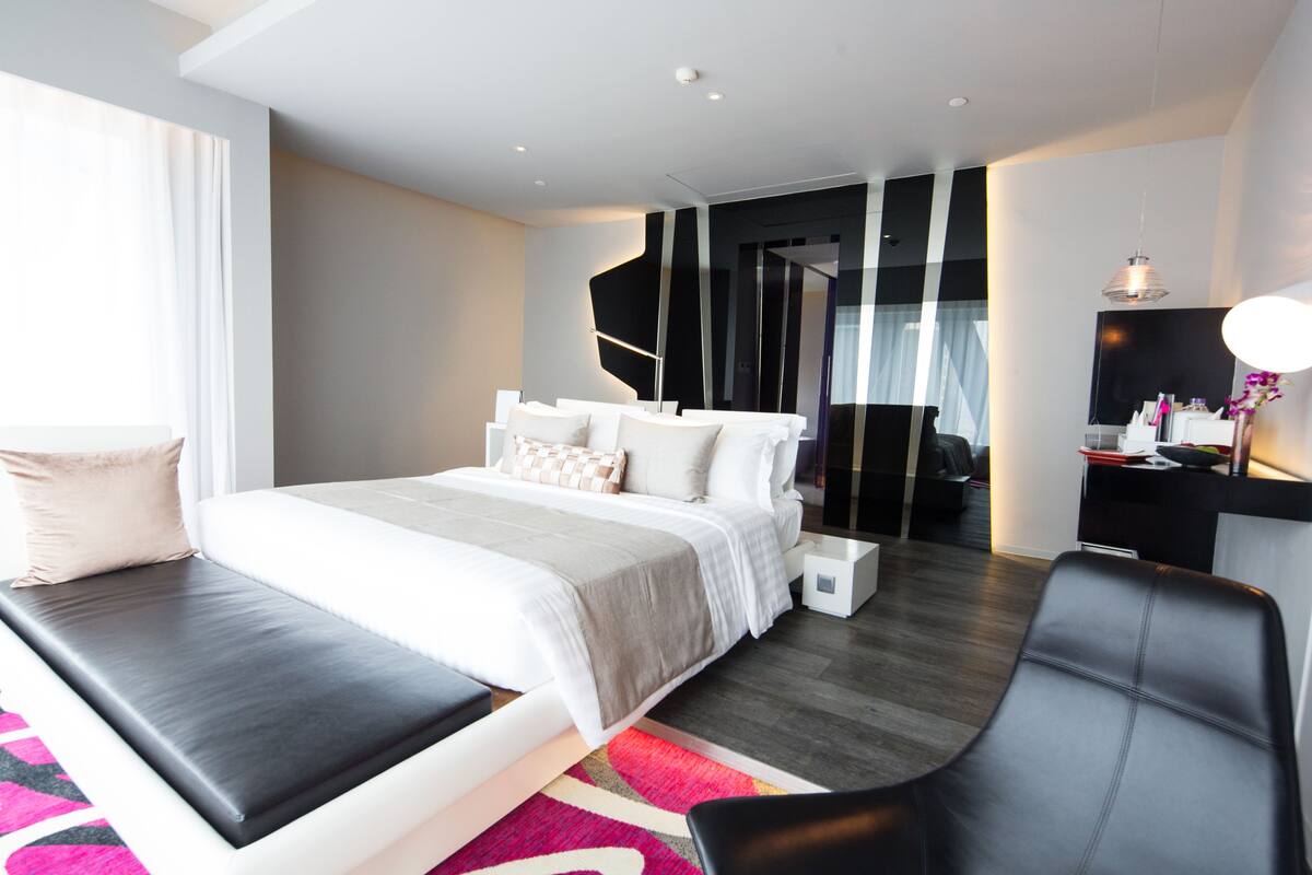A modern hotel room with a large bed, a seating area, a desk, and a mirror.