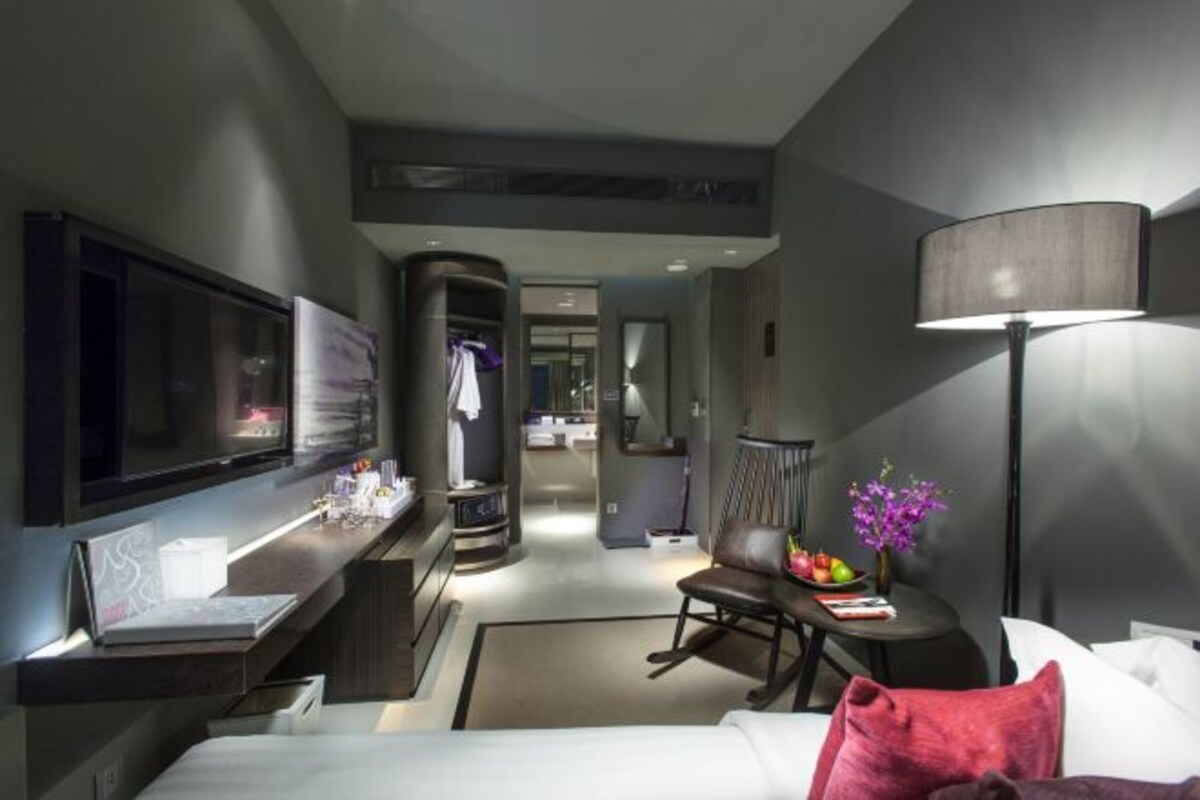 Una habitación de hotel moderna con una televisión de pantalla plana, un escritorio con lámpara, una silla y una cama con almohadas.