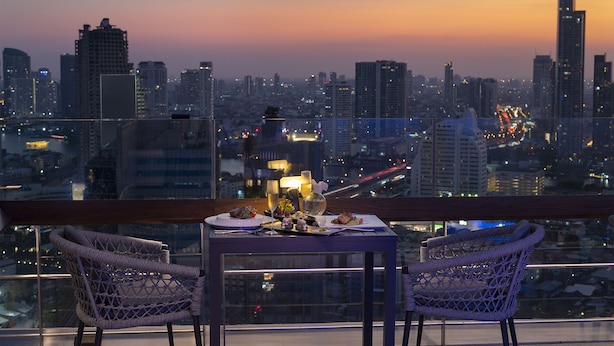 Un balcón con sillas de mimbre y una mesa dispuesta para comer, con vistas a un paisaje urbano al anochecer.