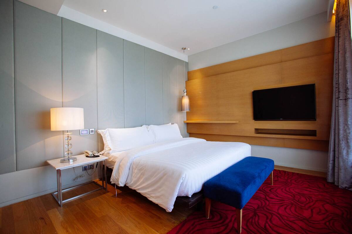 A hotel room with a bed, bedside table, lamp, and a TV mounted on the wall.