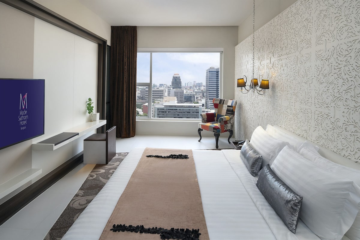 A modern hotel room with a large window, a flat-screen TV, a sofa, and a decorative rug.