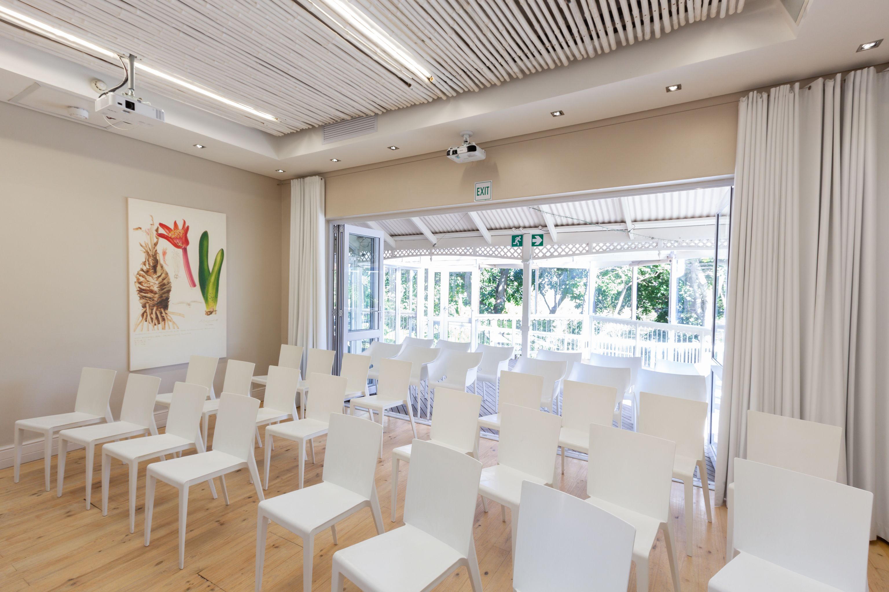 A conference room with white chairs arranged in rows, a projector on the ceiling, and a large window with curtains.