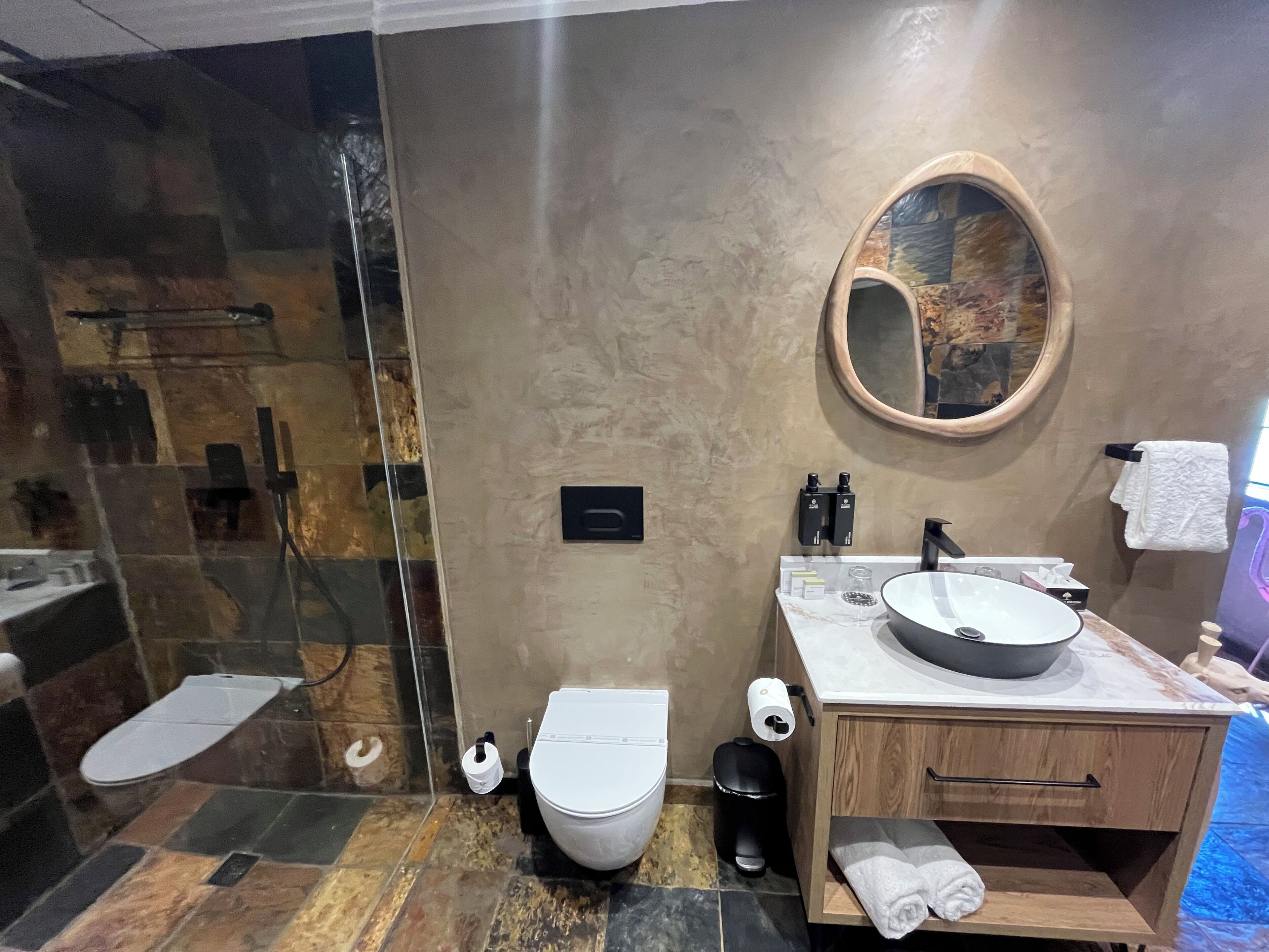 A modern bathroom with a glass shower enclosure, a toilet, a sink with a round mirror, and a wooden vanity.