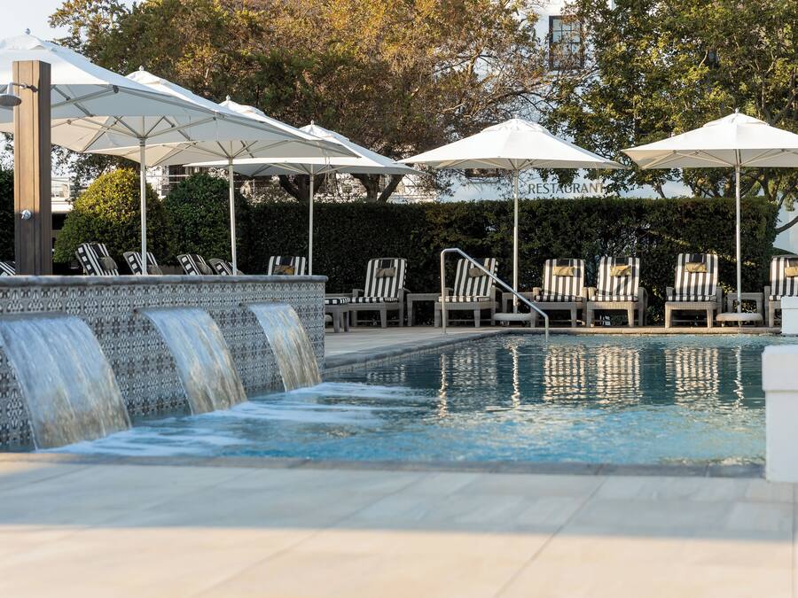 A pool area with multiple lounge chairs and umbrellas, a water feature, and a view of a restaurant in the background.
