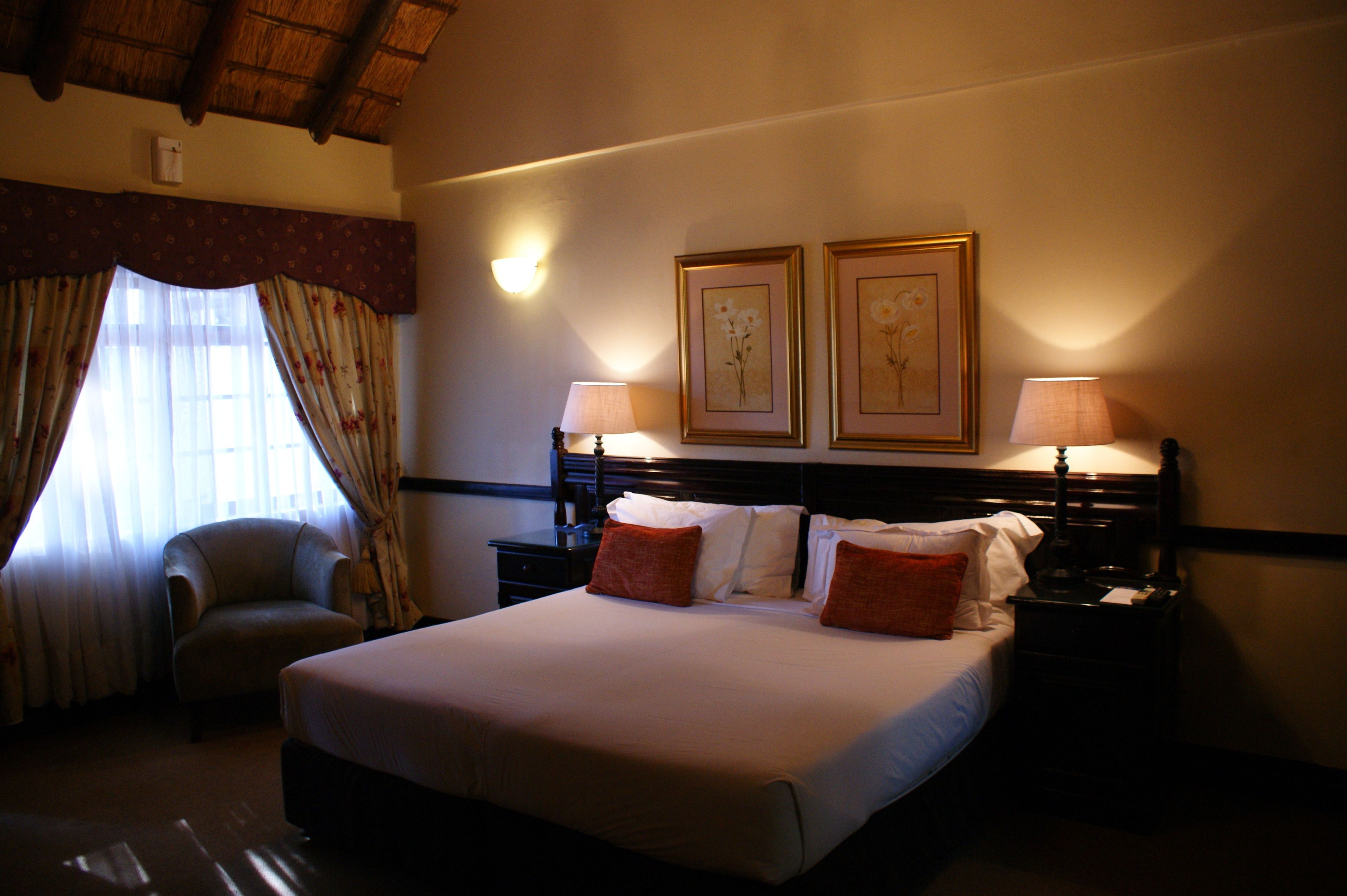 A hotel room with a large bed, two bedside lamps, a chair, and two framed floral paintings on the wall.