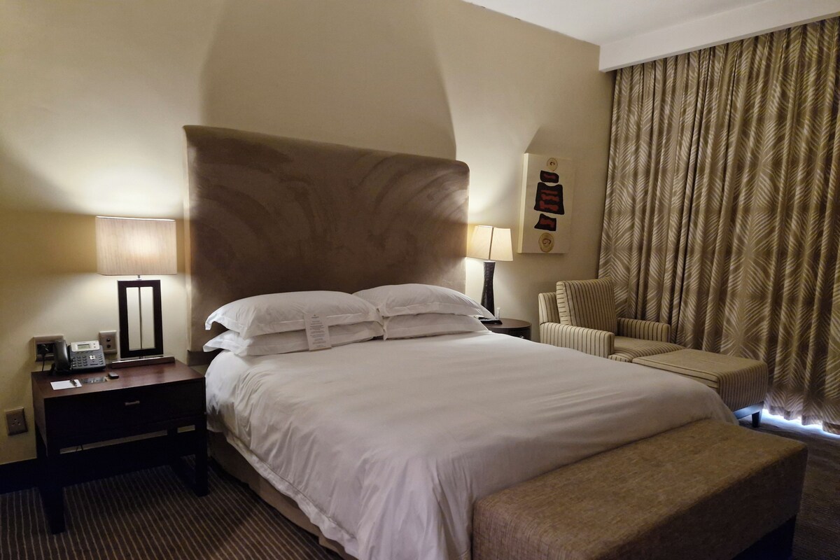 Ein Hotelzimmer mit einem großen Bett, einem Schreibtisch mit Telefon, einem Sessel und einem großen Fenster mit Vorhängen.