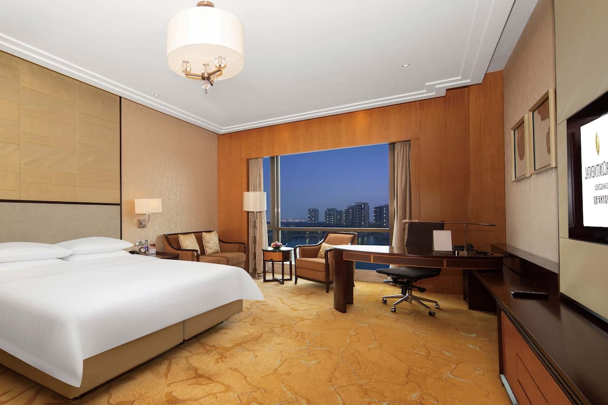Habitación de hotel con una cama grande, un escritorio, una silla, una lámpara y vista a la ciudad de noche.