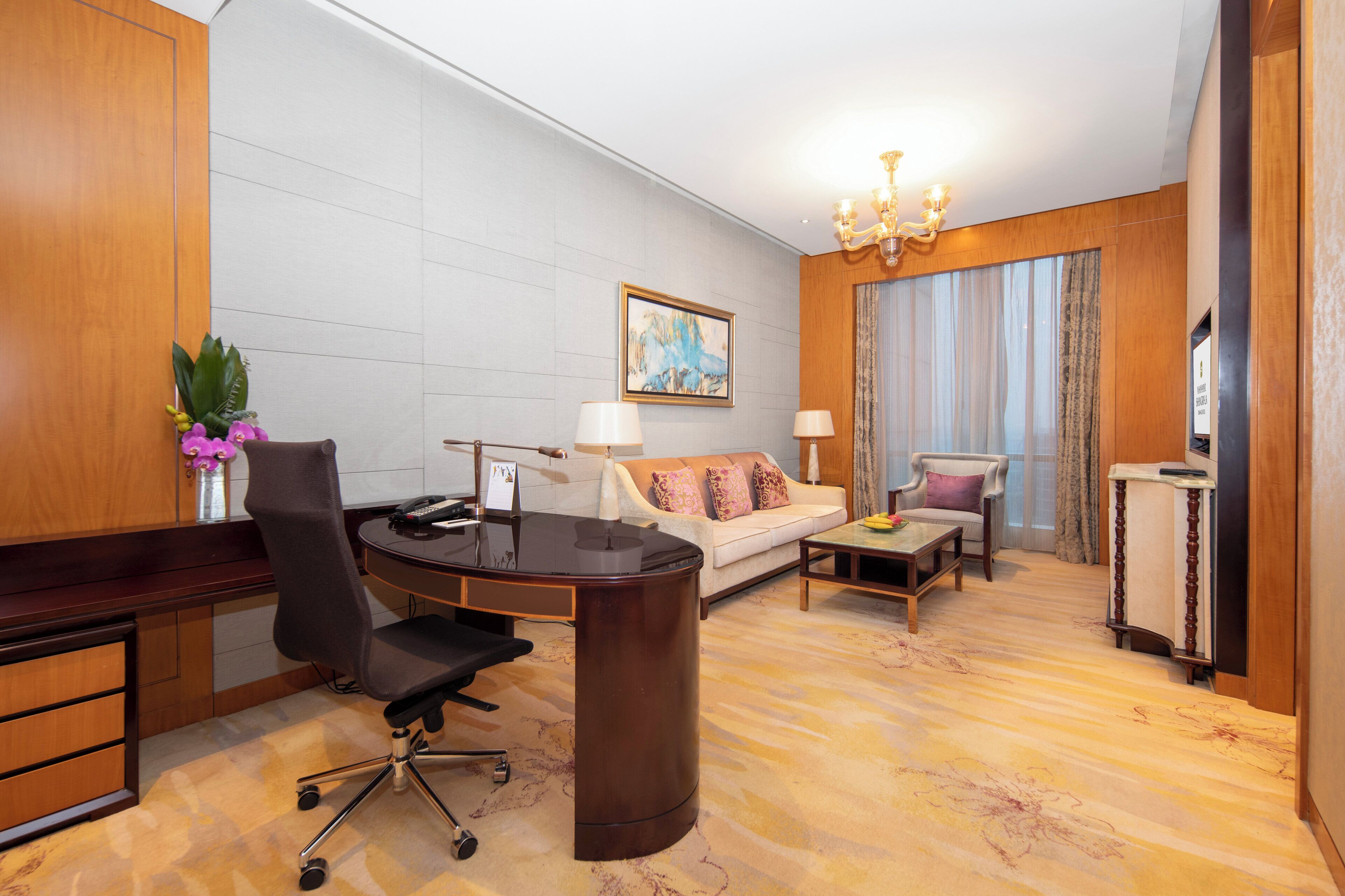 Ein Hotelzimmer mit einem Holztisch, einem schwarzen Bürostuhl, einem weißen Sofa mit gemusterten Kissen, einem kleinen Holztisch und einer Kristallleuchtre.