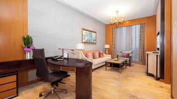 Ein Hotelzimmer mit einem Holztisch, einem schwarzen Bürostuhl, einem weißen Sofa mit gemusterten Kissen, einem kleinen Holztisch und einer Kristallleuchtre.